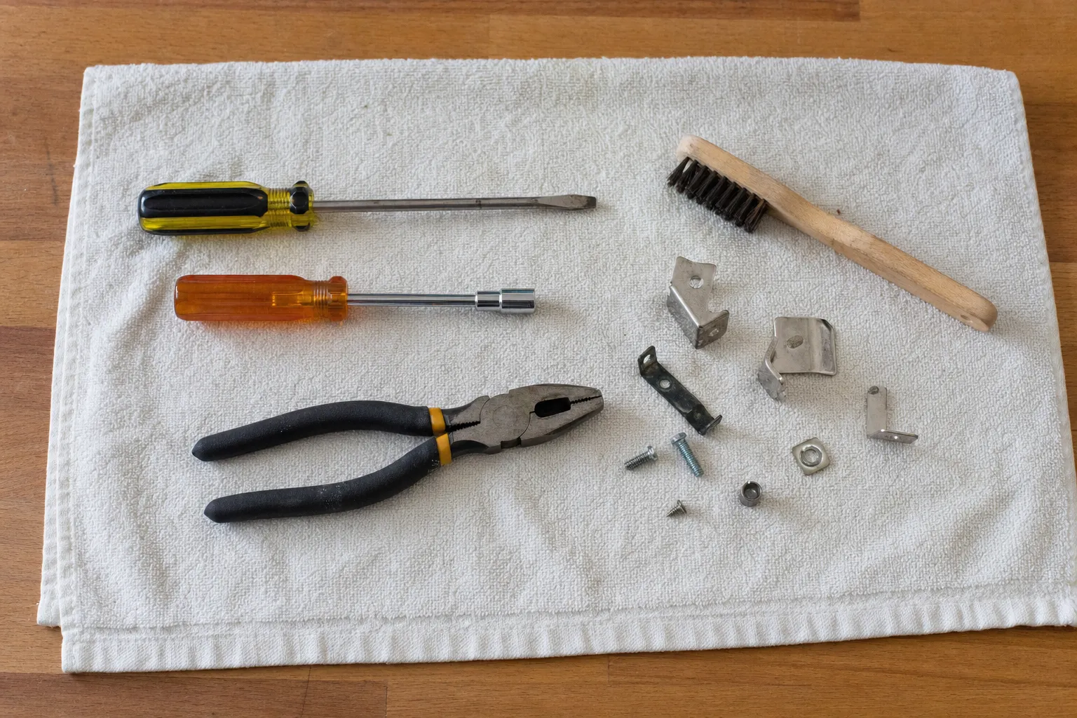 Flat screwdriver, orange nut driver, pliers and a wooden wire brush on a white towel with angle brackets, a clip, screws and a square nut