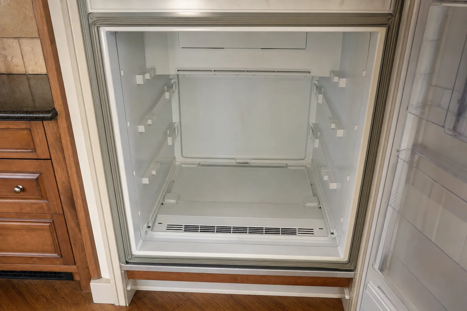 Built-in refrigerator compartment standing empty, shelf supports on the side walls, vent grille at the floor and the door gasket round the opening